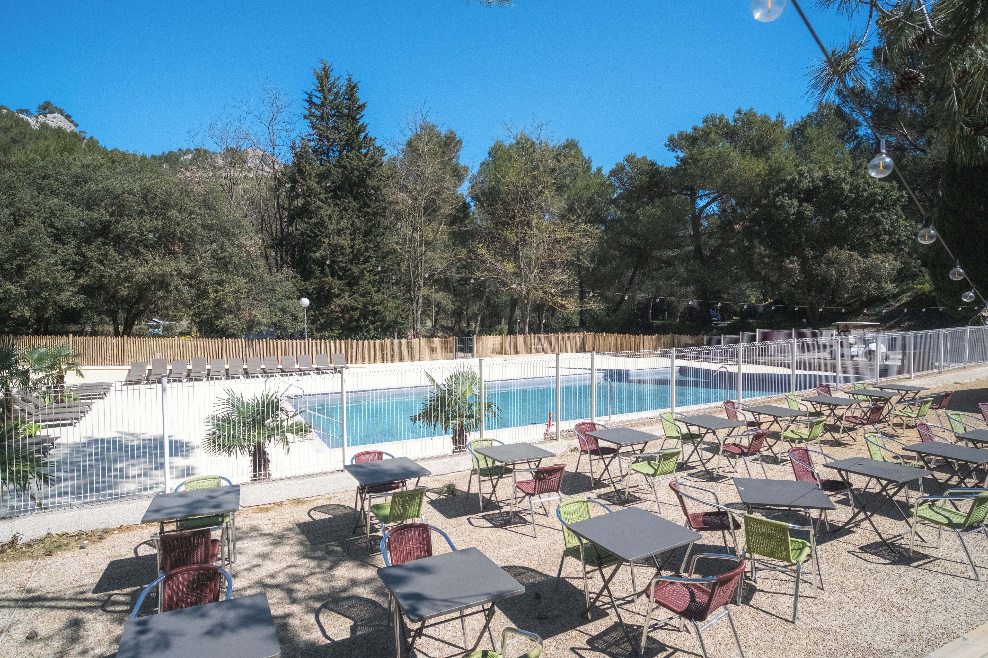 Zona de piscina al aire libre con mesas y sillas, rodeada por una valla y árboles.