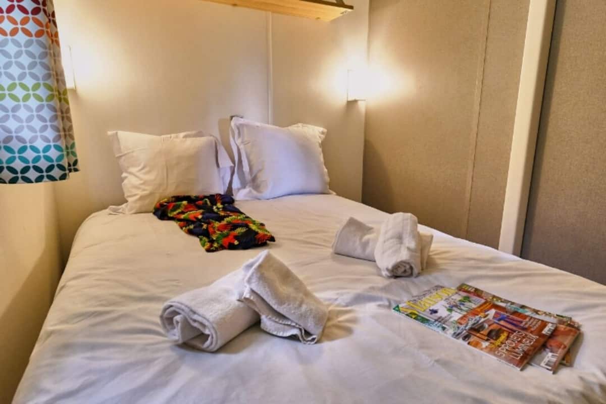A hotel room with a neatly made bed, white pillows, a colorful blanket, rolled towels, and a magazine on the bed.