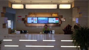 Una recepción con un mostrador, una pantalla digital mostrando información, un cartel que dice "GreenTree Inn" y una recepcionista.