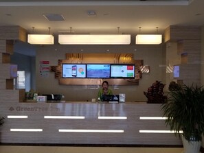 Una recepción con un mostrador, una pantalla digital mostrando información, un cartel que dice "GreenTree Inn" y una recepcionista.