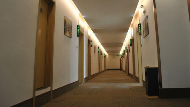 Pasillo de hotel con puertas señalizadas con letreros verdes, suelo alfombrado e iluminación empotrada en el techo.
