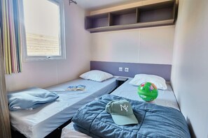 A dormitory room with two single beds, a window with blinds, a shelf above the beds, and a green helmet on one bed. - Ludo Camping Parc (Lussas)