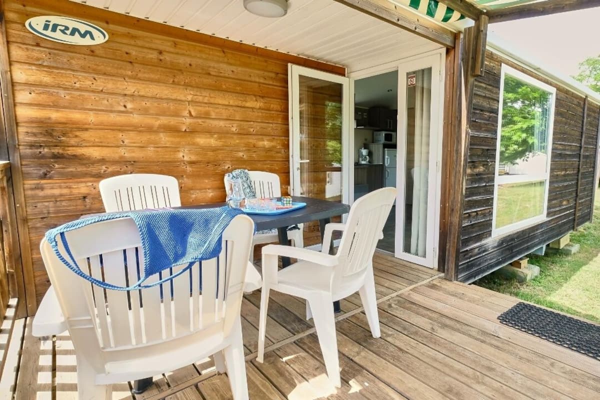 Una terraza de madera con una mesa y sillas, un área techada con un logotipo y una puerta corrediza de vidrio que da a una cocina.