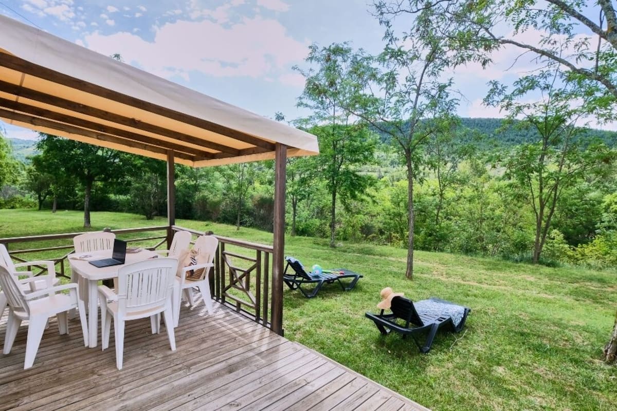 Un espacio al aire libre techado con una terraza de madera, sillas blancas y una mesa con un ordenador portátil.