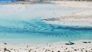 En strand med kristallklart, turkosfärgat vatten, en stenig kust och en båt som ligger förtöjd nära stranden.