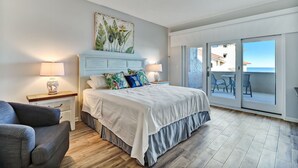 Un dormitorio con cama, mesitas de noche, una silla, un balcón con vistas y un cuadro en la pared.