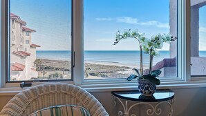 Una habitación con un gran ventanal que da a una playa y un edificio. Hay una silla de mimbre y una mesita con una orquídea en maceta.