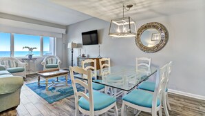 Una sala de estar moderna con una mesa de comedor de vidrio, sillas blancas y un ventanal con vistas al océano.