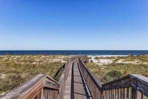 Ein hölzerner Steg führt zu einem Strand mit Dünen und dem Meer im Hintergrund.