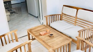 Un salon accueillant avec un canapé, deux fauteuils en bois et une petite table sur laquelle se trouve un plateau. On aperçoit une cuisine au fond.