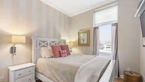 Ein ordentlich eingerichtetes Schlafzimmer mit Bett, Nachttischen, einem Fenster mit Vorhängen und einem gerahmten Bild an der Wand.