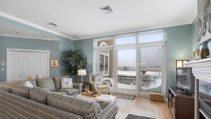 Ein Wohnzimmer mit einer großen Couch, einem Fernseher und einem Kamin. Ein großes Fenster bietet Blick ins Freie.
