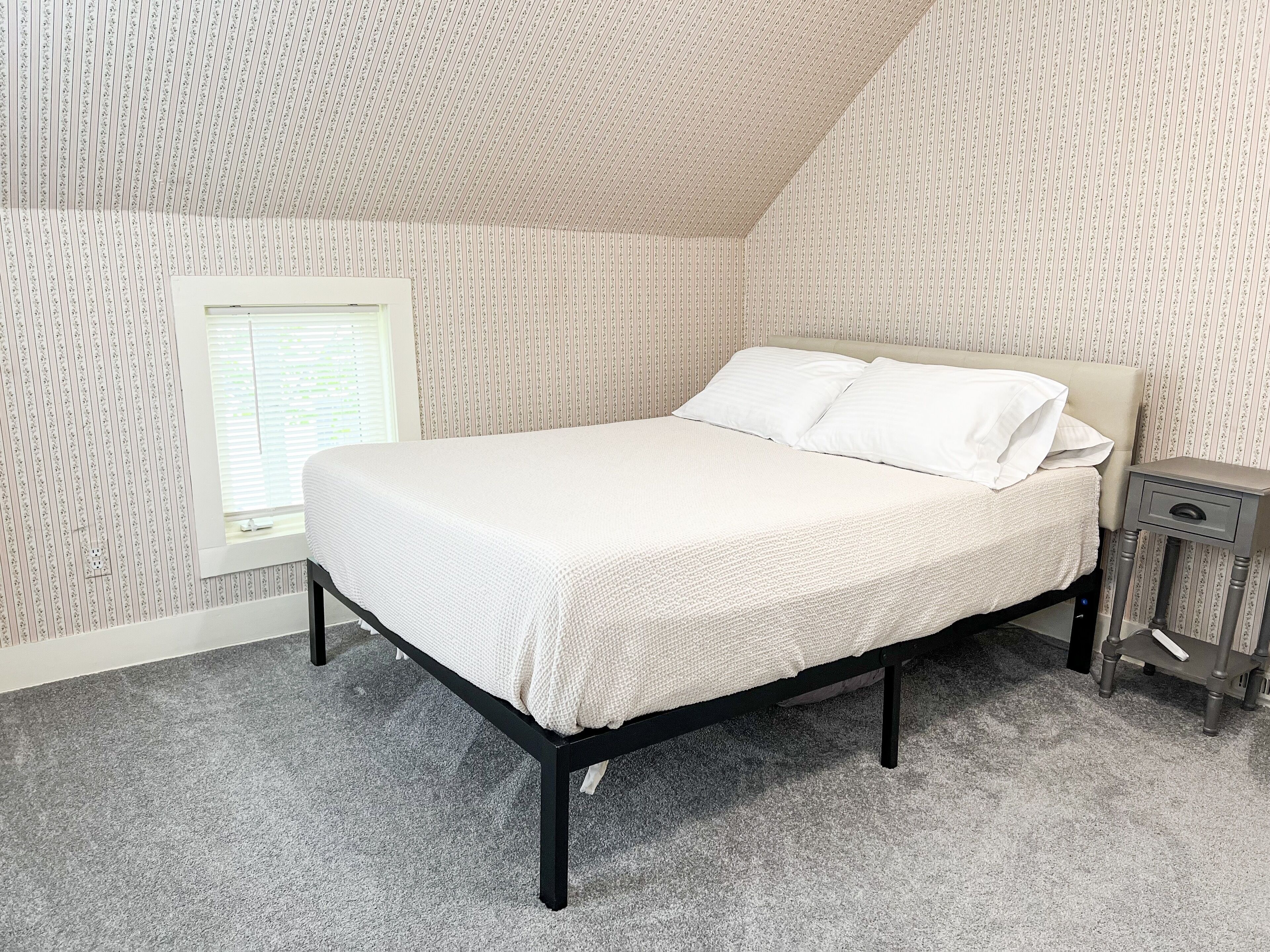 A bedroom with a bed, a nightstand, a window, and striped wallpaper.
