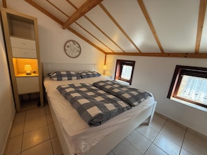 Ein Schlafzimmer mit Bett, Nachttischen, einem Fenster und einer Holzbalkendecke.