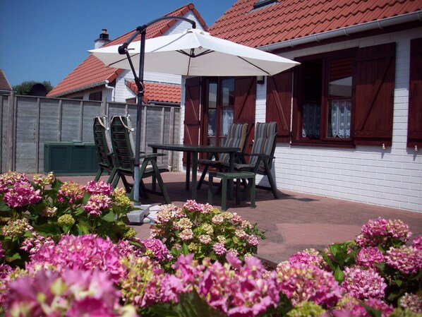 A patio with a table and chairs, surrounded by pink flowers.