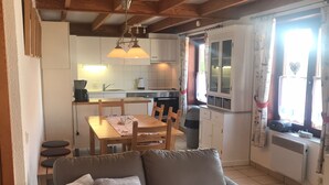 Uma sala de estar acolhedora com sofá e mesa de jantar, uma cozinha com armários brancos e uma janela, e um ventilador de teto.