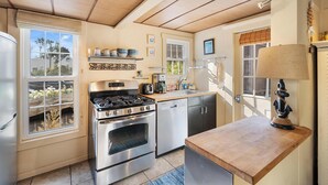 A kitchen with a gas stove, oven, dishwasher, and wooden countertop. There are windows with blinds, a hanging light fixture, and a small shelf with dishes.