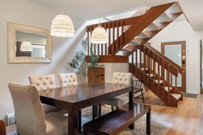 Un comedor con mesa y sillas de madera, una escalera e iluminación colgante.