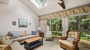 Ein Wohnzimmer mit einer Couch, einem Sessel und einem Couchtisch, ein großes Fenster mit Blick auf Bäume und ein Dachfenster.