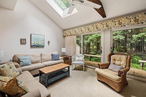 A living room with a sofa, armchair, and coffee table, a large window with a view of trees, and a skylight.