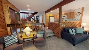 A cozy living room with a sofa, chairs, a lamp, and a staircase leading to a kitchen area. - Harbor Cove! 5 Bedroom! #82 Ski, Golf, Shop! (Harbor Springs)