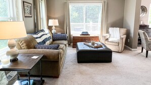 A cozy living room with a sofa, ottoman, and armchair. There is a table with a lamp and a tray with a mug. A window with curtains and a view of greenery is visible. - COMFORTABLE & SPACIOUS Home in Harbor Springs - Minutes from the Ski Hills!! (Harbor Springs)