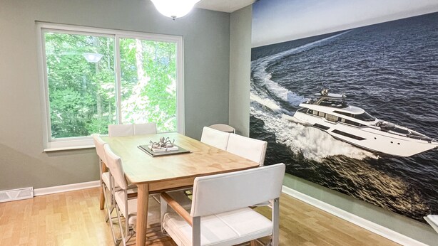 A dining area with a wooden table and white chairs, a window with a view of greenery, and a large mural of a yacht on the ocean. - COMFORTABLE & SPACIOUS Home in Harbor Springs - Minutes from the Ski Hills!! (Harbor Springs)