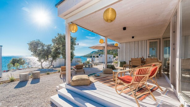 A wooden deck with outdoor furniture, a view of the sea, and a building with a glass door. - Buqez - 2BR - Beach Villa 42 (Pakostane)