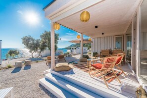 A wooden deck with outdoor furniture, a view of the sea, and a building with a glass door. - Buqez - 2BR - Beach Villa 42 (Pakostane)