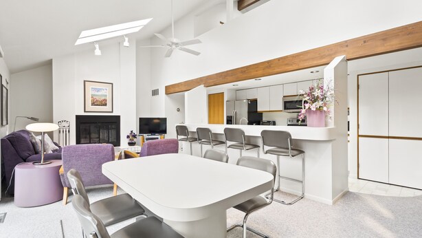 Un salon moderne avec une table à manger blanche, un canapé violet et un coin cuisine doté de tabourets de bar.
