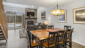 A dining area with a wooden table and chairs, a stone fireplace, and a large window with a view of the outdoors. - Trout Creek #122 SHUTTLE TO NUBS! POOLS &Fitness! (Harbor Springs)