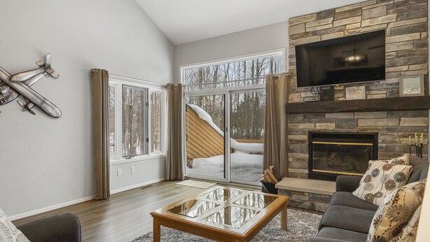A living room with a stone fireplace, a flat-screen TV, a glass coffee table, and a view of a snowy landscape through large windows. - Trout Creek #122 SHUTTLE TO NUBS! POOLS &Fitness! (Harbor Springs)
