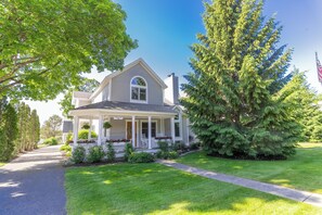 A two-story house with a covered porch, a front yard with a lawn, and a large evergreen tree. - Villa East - Main Street! 4BDR 3.5BATH - Sleeps 10! (Harbor Springs)