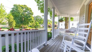 Une véranda en bois avec des balustrades blanches, une chaise berçante et un pot de fleurs suspendu.