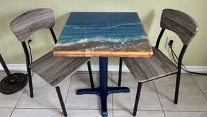 Une table dont le plateau est en verre et orné d’un motif représentant un paysage marin, flanquée de deux chaises à dossier en osier, le tout posé sur un sol carrelé devant un mur vert clair.