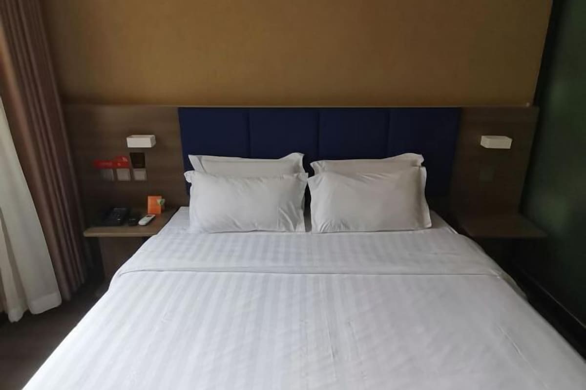 A hotel room with a bed, two pillows, a nightstand with a phone and a remote control, and a wall-mounted light.