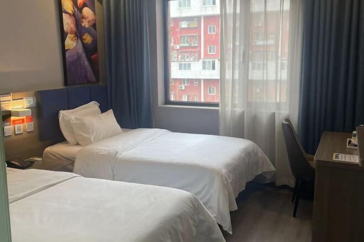 A hotel room with two beds, a desk, a chair, a painting on the wall, and a window with curtains.