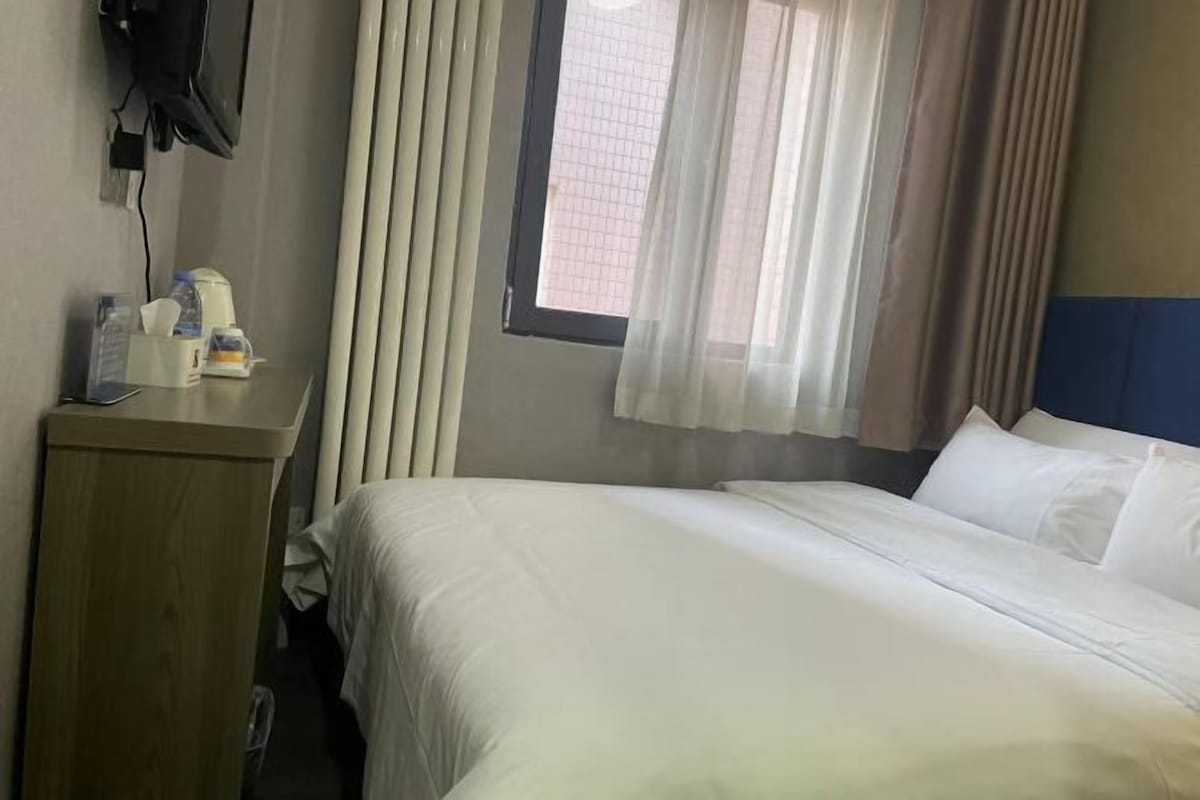 A hotel room with a bed, a desk with a telephone, a window with curtains, and a wall-mounted TV.