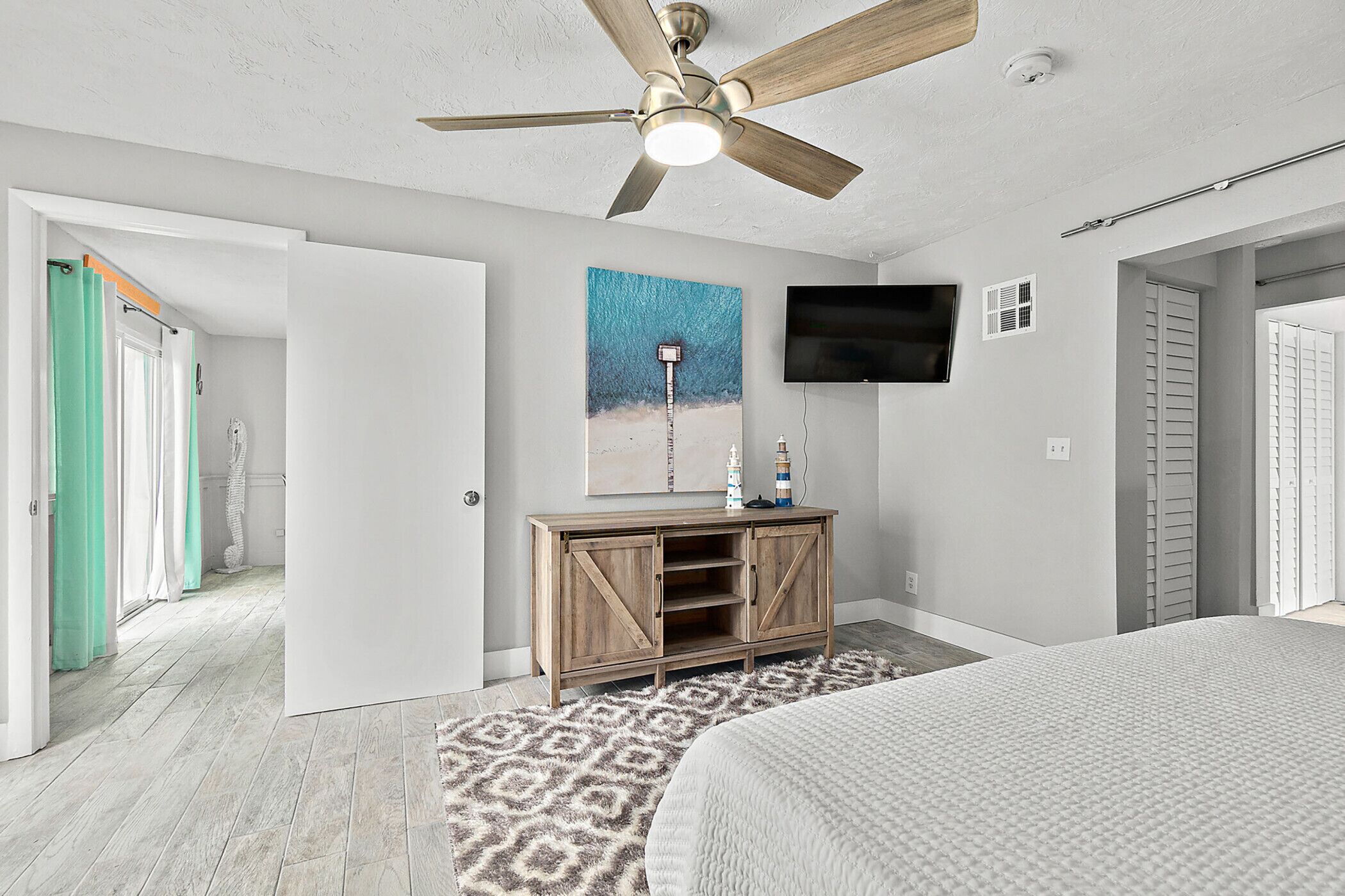 Quarto com cama, ventilador de teto, televisão presa na parede e cômoda de madeira.
