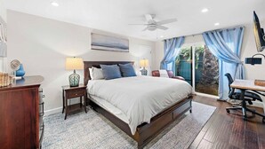 Un dormitorio con una cama grande, dos mesitas de noche con lámparas, un ventilador de techo y vistas a un jardín a través de un ventanal corredero.