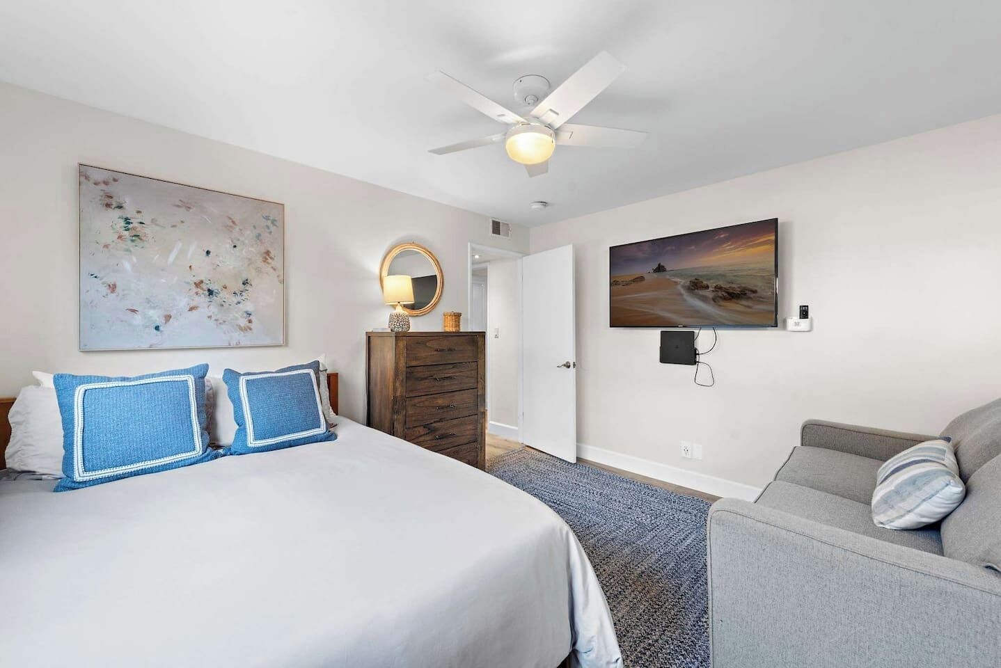 Quarto de hotel com cama, penteadeira, cadeira, ventilador de teto, quadro e televisão.