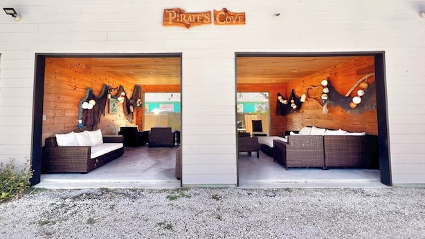 A wooden cabin with two large doors labeled "Pirate's Cove." Inside, there are wicker sofas and a small table. - Sunshine Shack - 2 bedroom, In-Town, Dog Friendly, Near the Beach! (Port Aransas)