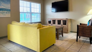 A living room with a yellow sofa, a wooden cabinet, a television, and a ceiling fan. - Sunshine Shack - 2 bedroom, In-Town, Dog Friendly, Near the Beach! (Port Aransas)