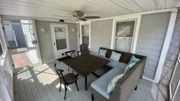 Un patio couvert avec une table et des chaises en bois, un canapé en osier agrémenté de coussins à motifs et un ventilateur de plafond.