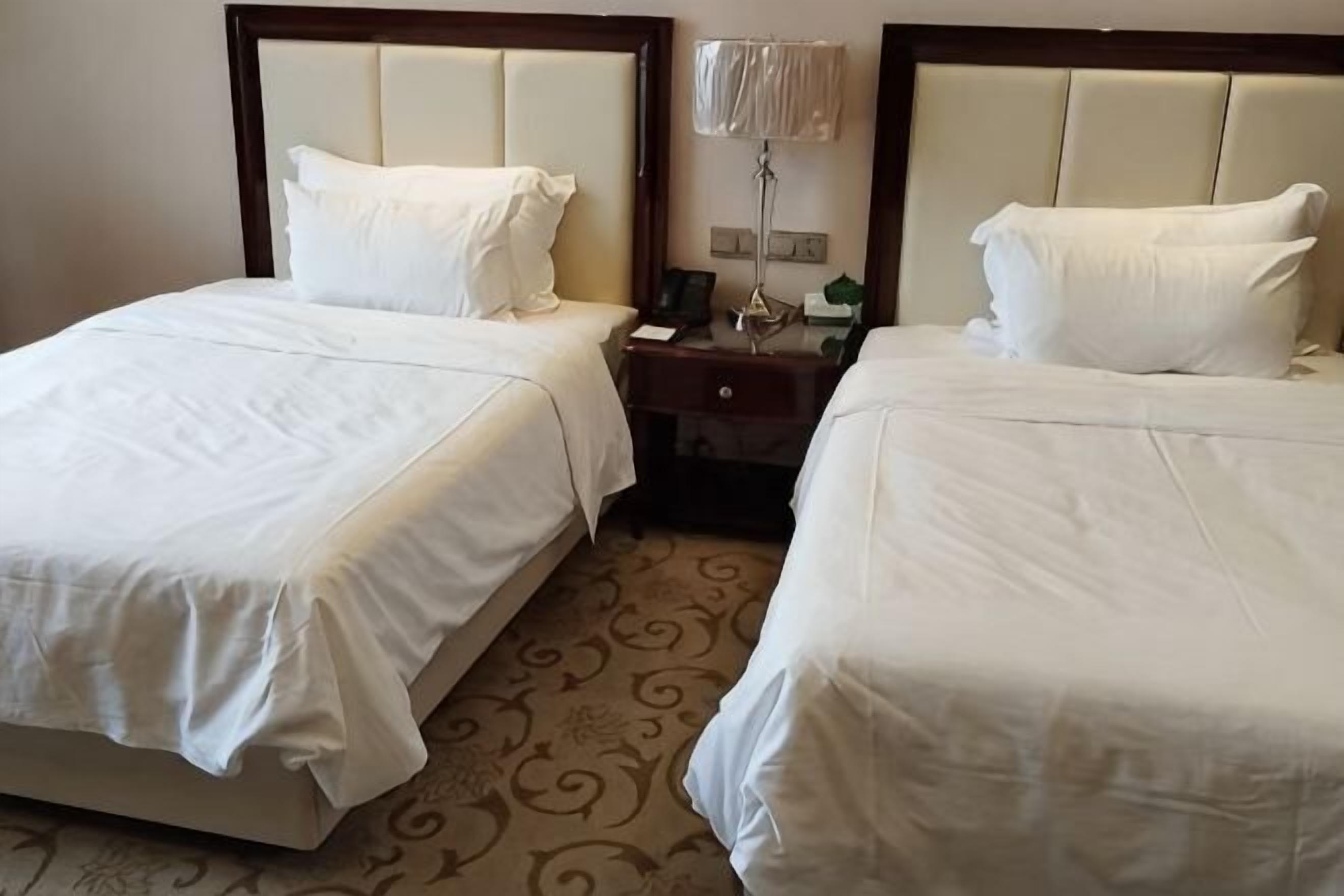 Dos camas con sábanas blancas, una mesita de noche con lámpara y una alfombra estampada.