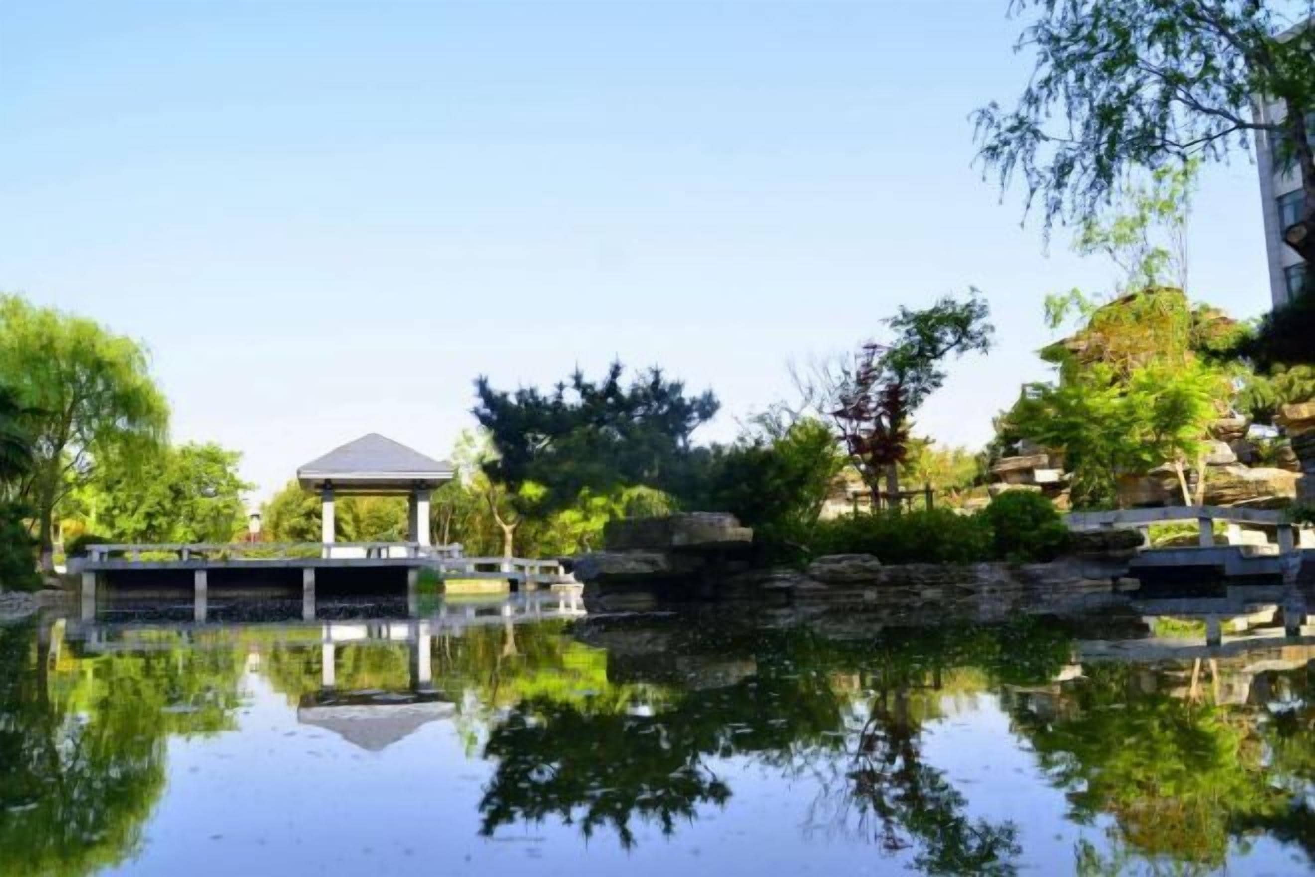 A tranquil outdoor setting with a pavilion, a calm water body reflecting the surroundings, and a stone bridge.