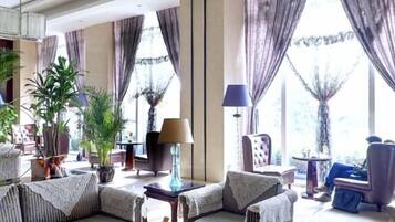 A well-lit living room with a sofa, armchairs, a coffee table, and a lamp. There are large windows with curtains, and potted plants are visible.