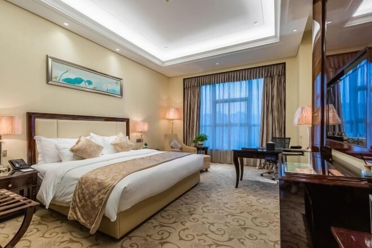Una habitación de hotel con una cama grande, un escritorio con computadora, una silla, una lámpara y un cuadro en la pared.