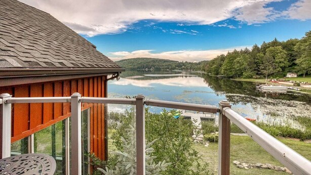 Een houten hutje met een balkon met uitzicht op een kalm meer, omgeven door weelderige begroeiing en heuvels.