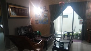 Una sala de estar con un sofá de madera, una mesa de centro de vidrio y una mesa de comedor con sillas. Hay cuadros en la pared y una ventana con cortinas.